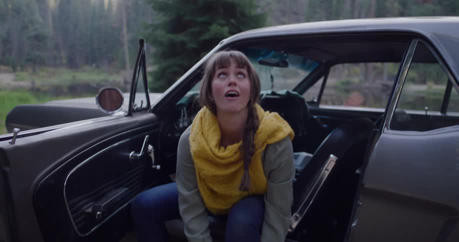 A young woman in a classic car looks up at the sky in wonder before tying her hiking boots for an adventure in the woods