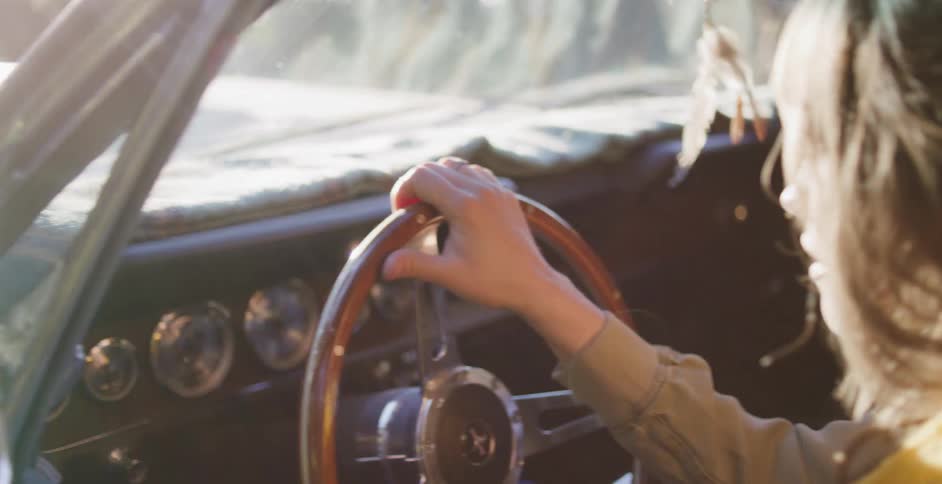 A woman drives a classic car down a scenic road, with sunlight flaring through the windshield