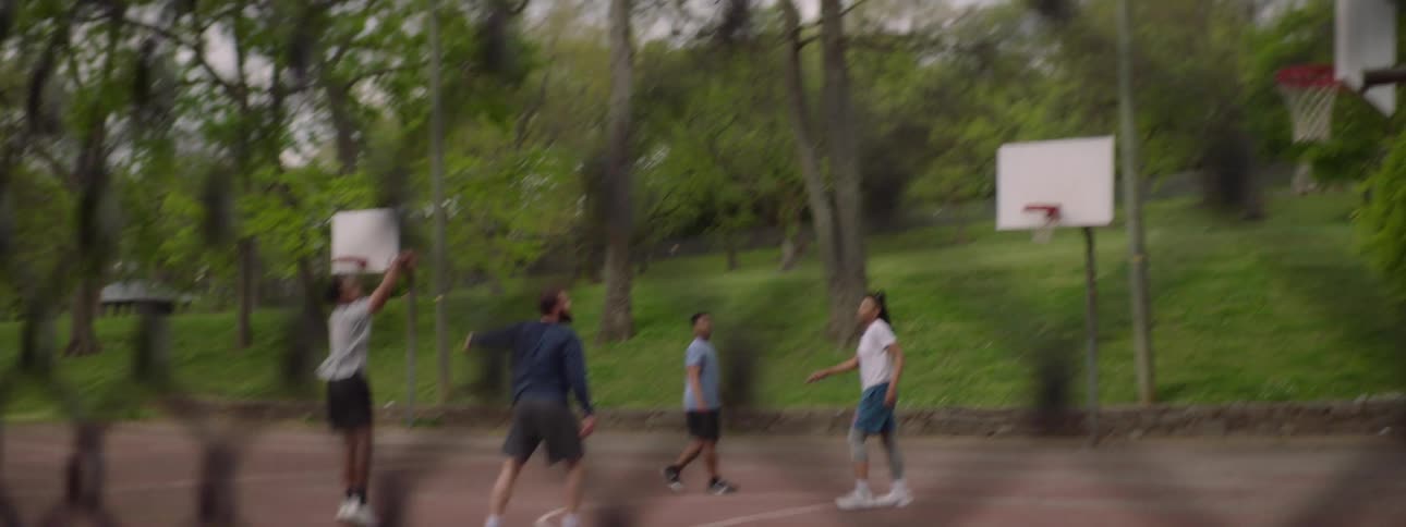 A man sinks a jump shot in slow motion during a pickup basketball game with friends at an outdoor court, viewed from behind a fence