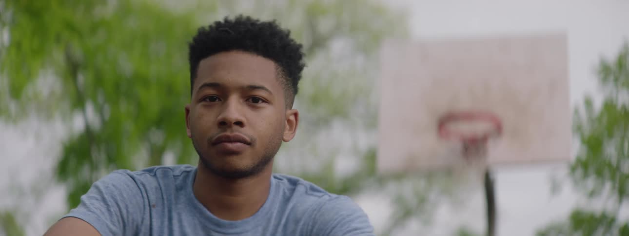 Portrait of a serious young man sitting in front of a basketball hoop on an outdoor court