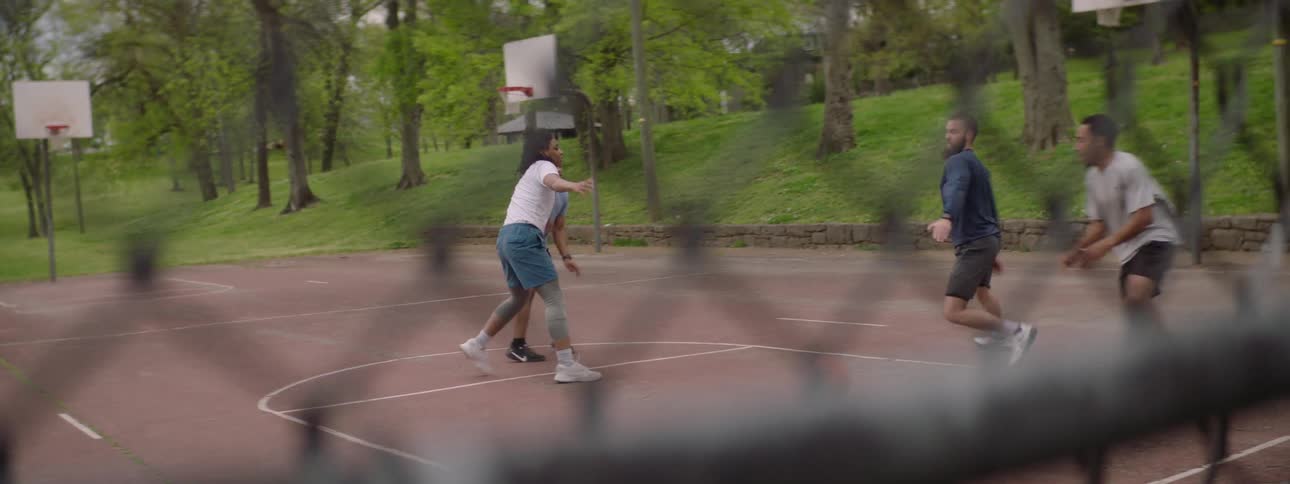 Friends play a pickup basketball game at a park in slow motion, seen through a chain-link fence, ending with a successful layup