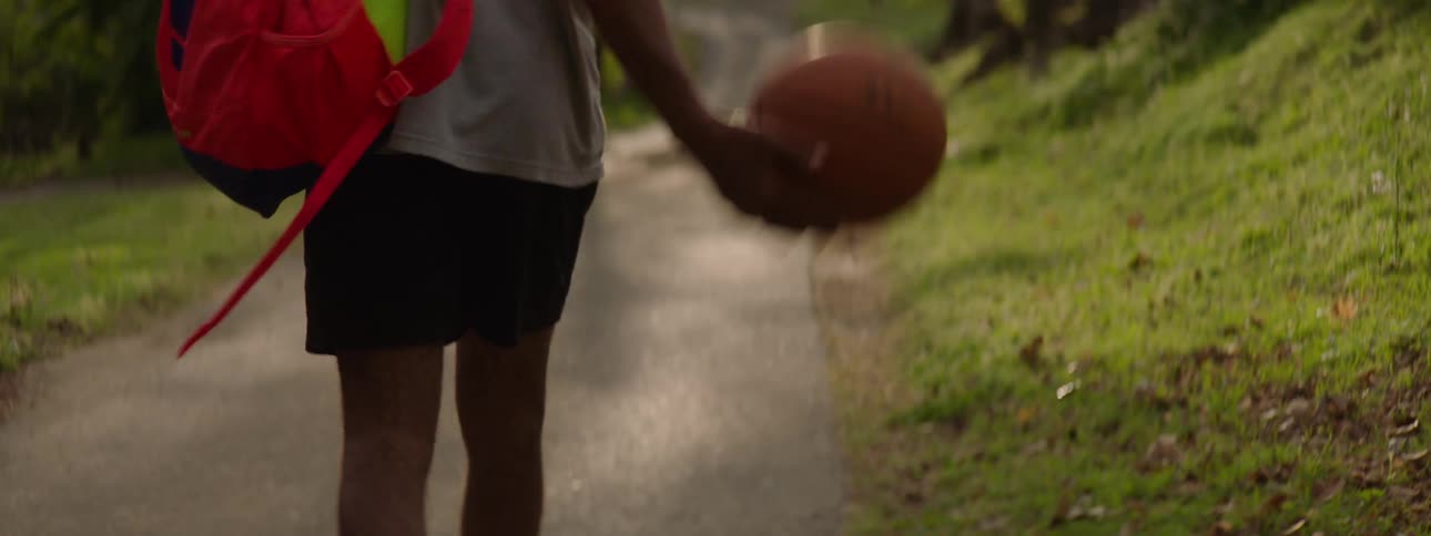 Slow motion from behind shot of a young basketball player walking down a sunlit park path while carrying a backpack and dribbling a ball