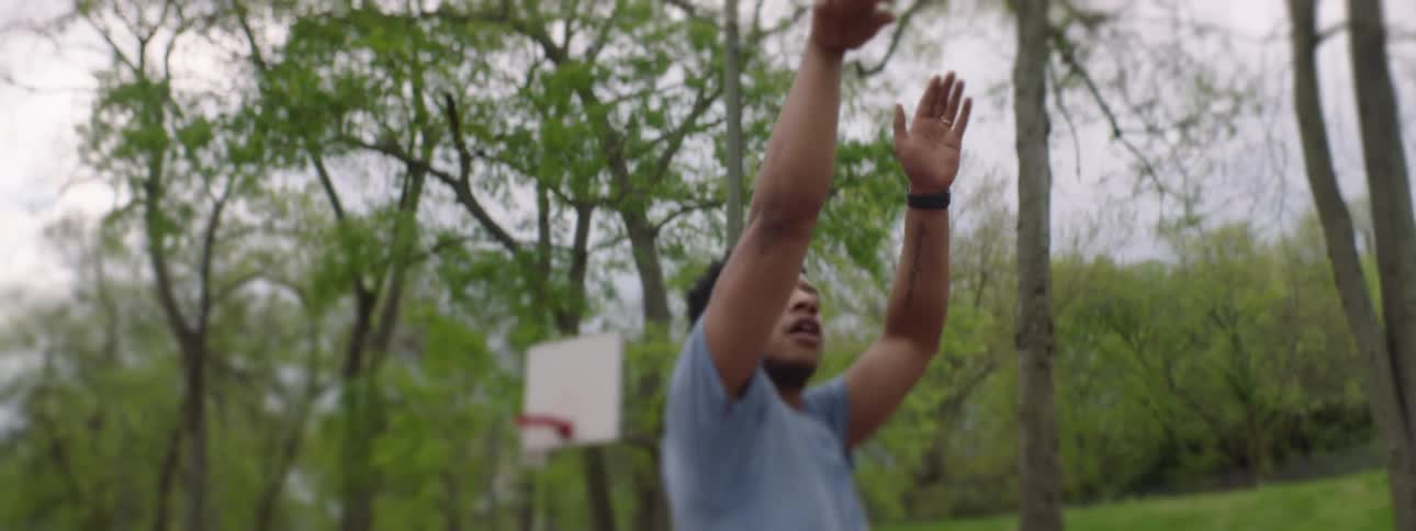A young man shoots a basketball in slow motion on an outdoor court surrounded by lush green trees