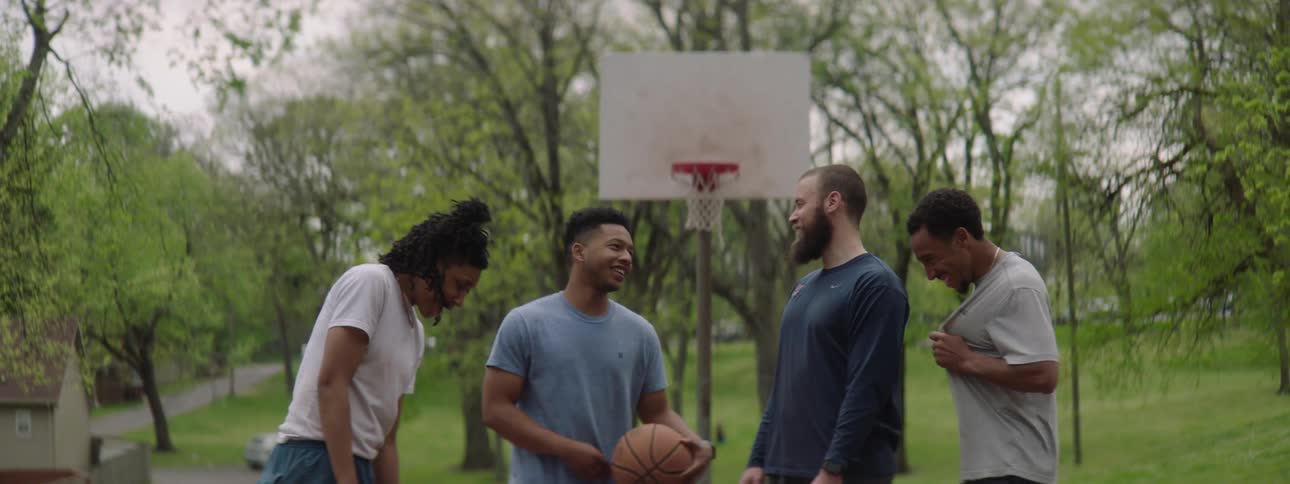 A diverse group of four friends laugh and talk together in slow motion on an outdoor basketball court in a park