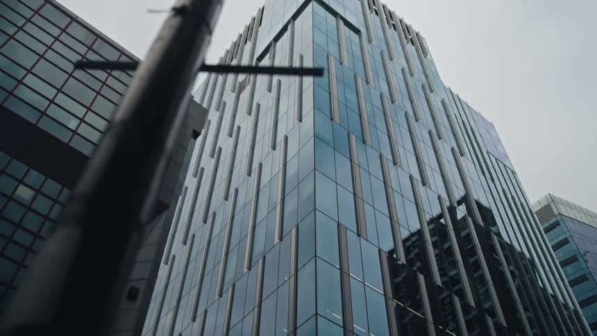 A slow, low-angle reveal of a modern glass skyscraper reflecting the city against a grey, overcast sky