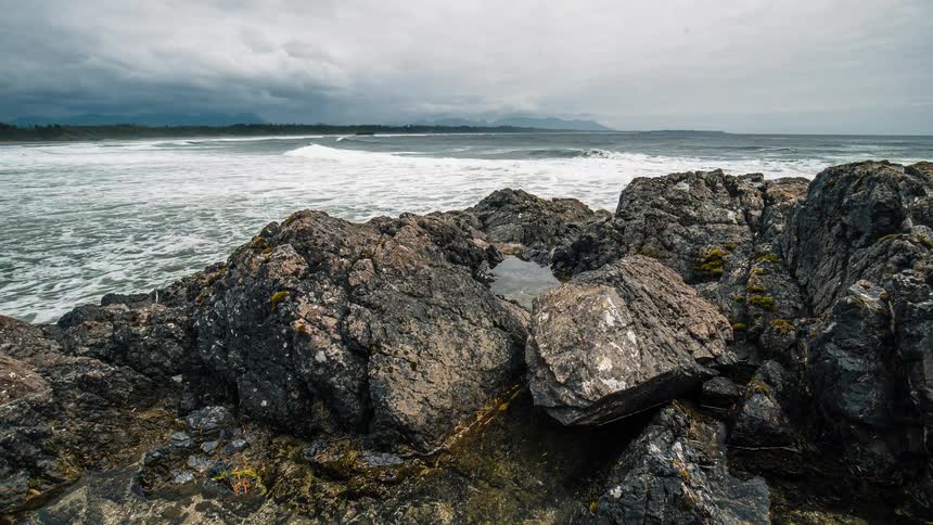 A dramatic timelapse zooms out from a rocky tide pool to reveal a vast, moody ocean with crashing waves under a cloudy sky