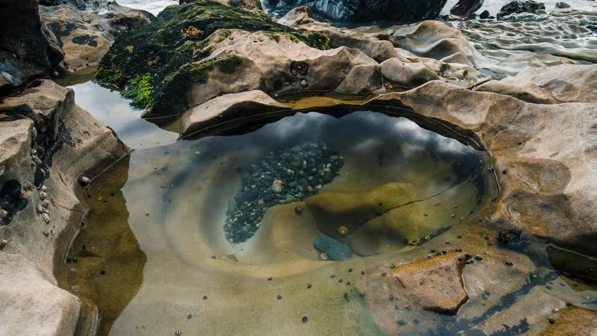 A tranquil timelapse zooms into a tide pool on a rocky shore, its still water perfectly reflecting the passing clouds