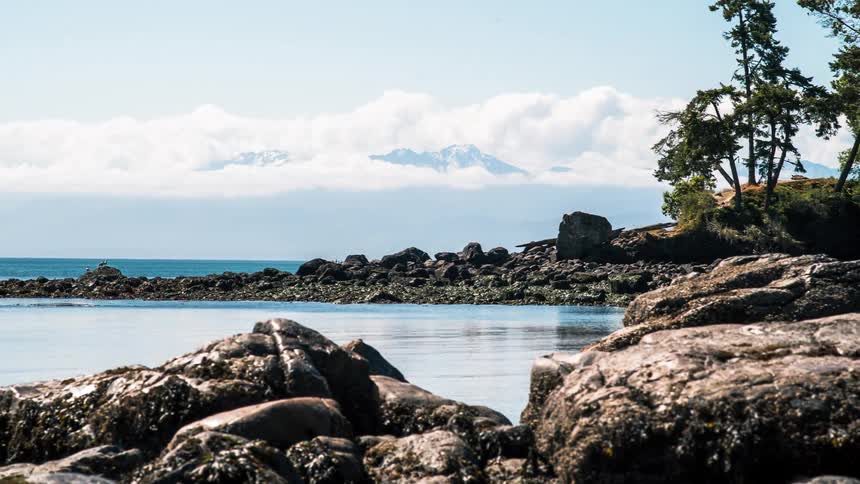 A serene timelapse from a rocky shore looking out over the water towards distant snow-capped mountains as clouds drift by
