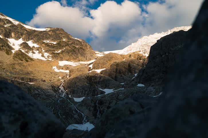 A dramatic dolly shot reveals a sun-kissed mountain glacier with meltwater streams flowing down the rocky valley