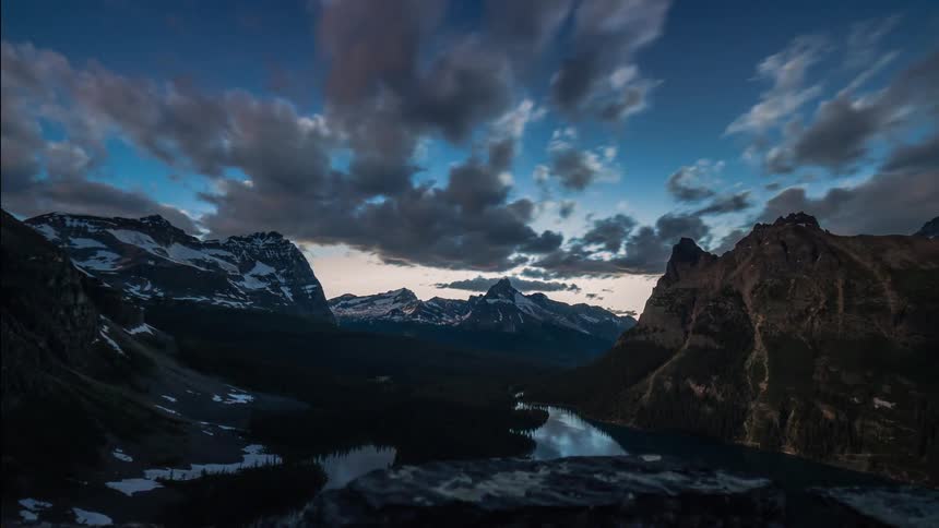 A dramatic time-lapse reveals a beautiful sunrise over a snow-capped mountain range and pristine lake as clouds roll through the valley