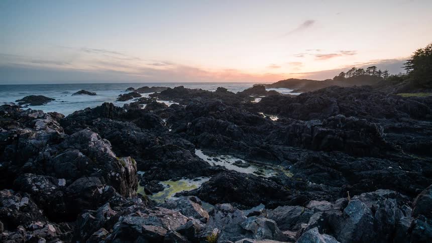 A dramatic time-lapse reveals ocean waves crashing against a rugged, black volcanic rock shoreline during a beautiful sunset
