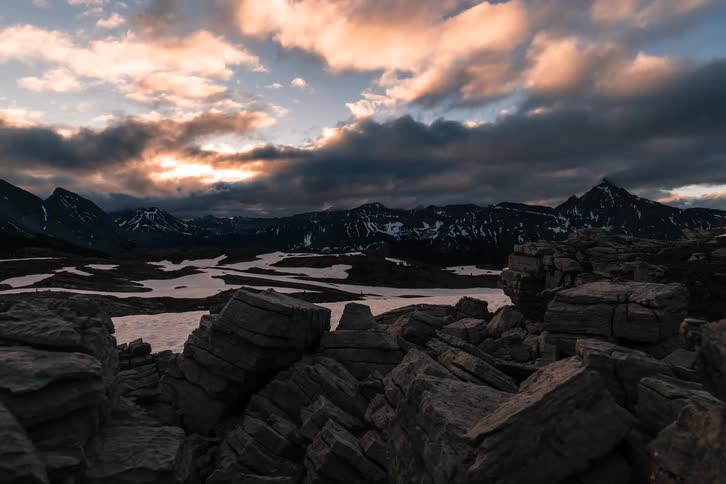 An epic time-lapse captures the sunrise illuminating dramatic clouds over a desolate, rocky, and snowy mountain range