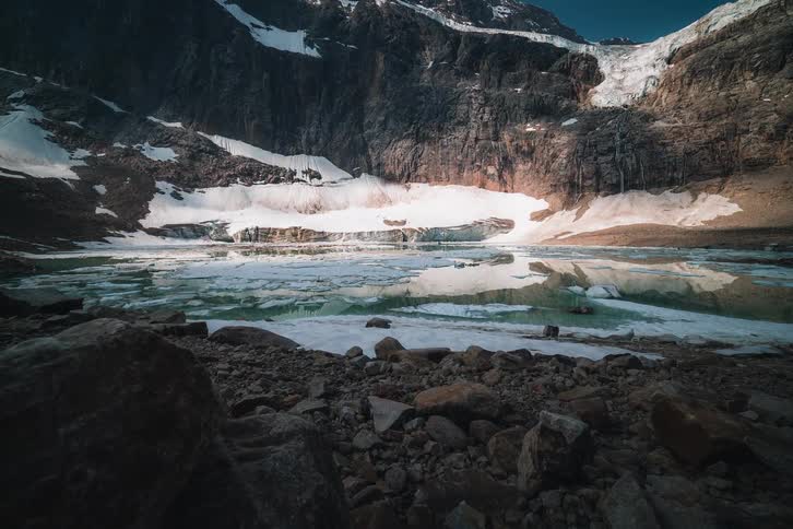 A dolly timelapse shows shadows moving across a majestic, snow-covered mountain and a partially frozen glacial lake with turquoise water
