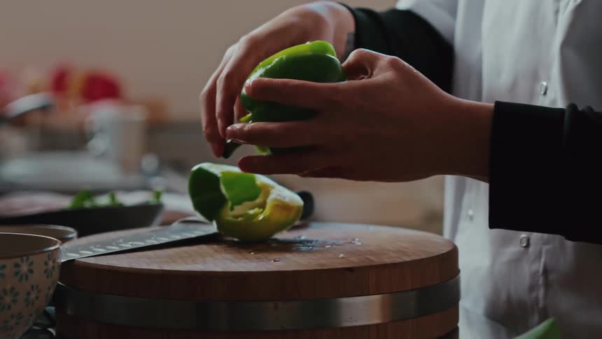 A chefs hands carefully slice a fresh green bell pepper in half and remove the seeds on a round wooden cutting board in slow motion