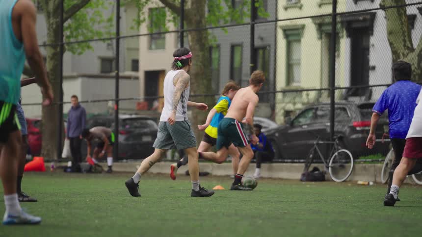 A diverse group of young adults enjoys a casual game of soccer on a turf field in a city park in slow motion