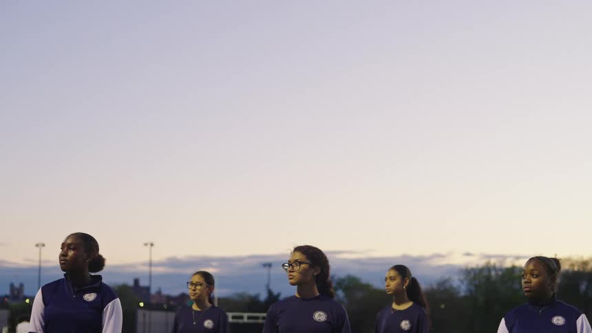 A diverse team of young women stands united and focused on a sports field at twilight