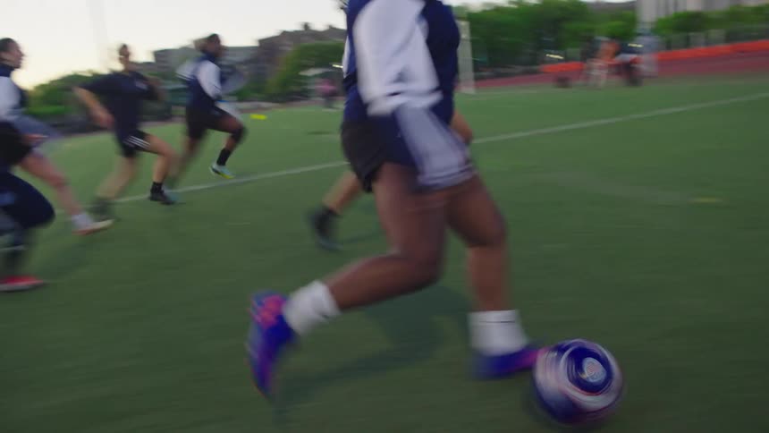 A slow-motion tracking shot follows a young female soccer player dribbling a ball down the field as her teammates run alongside her during an evening practice