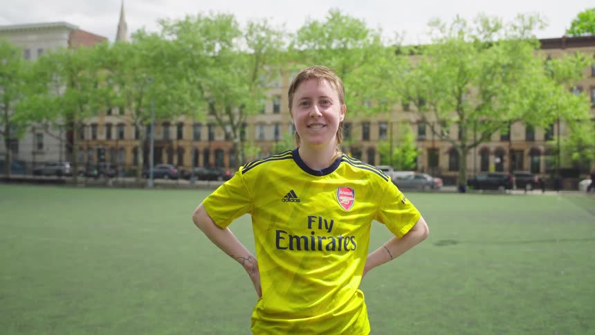 A confident female soccer player in a yellow jersey smiles proudly on an urban soccer field