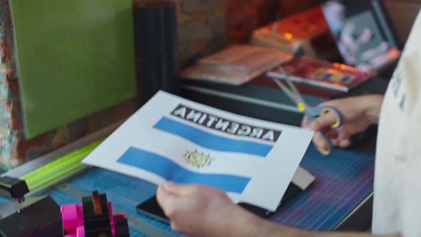 A person cuts out an Argentina flag design and places it on a white t-shirt in a heat press for custom printing