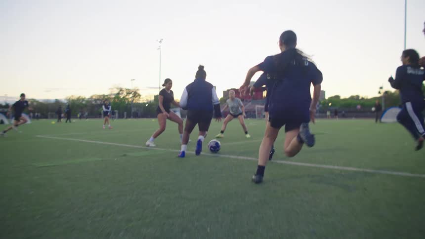 A slow-motion, low-angle shot follows a female soccer player as she dribbles the ball down the field, with teammates defending during an evening practice