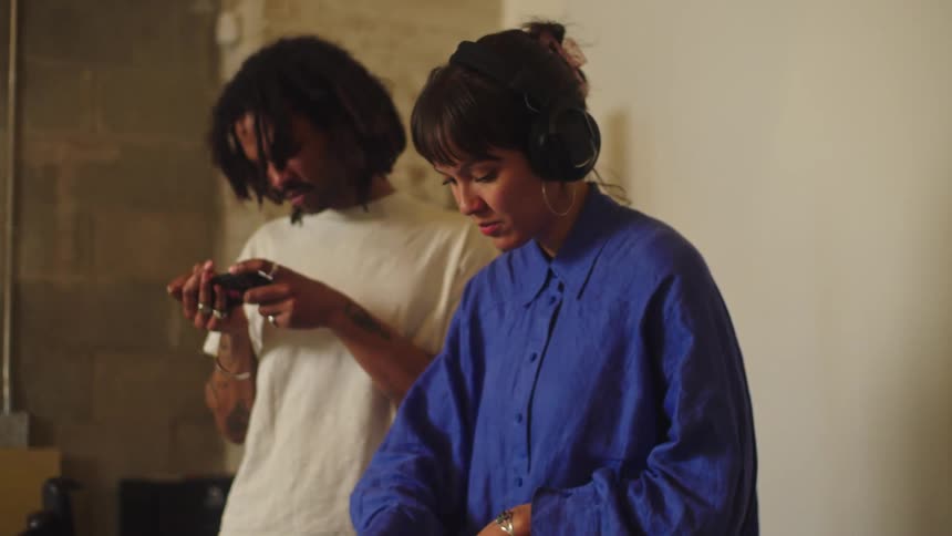 A female DJ wearing headphones intently mixes music on her deck while a man stands beside her using his phone