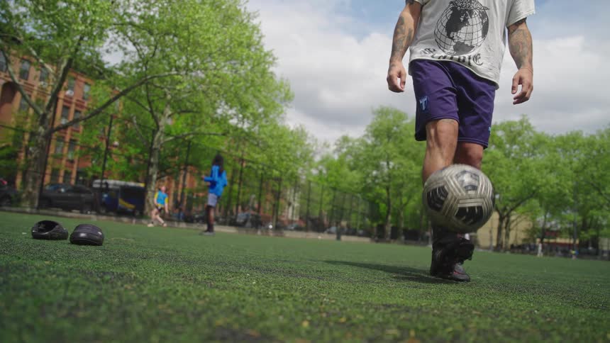 In stunning slow motion, a soccer player skillfully balances a ball on his foot before kicking it up on a city park turf field