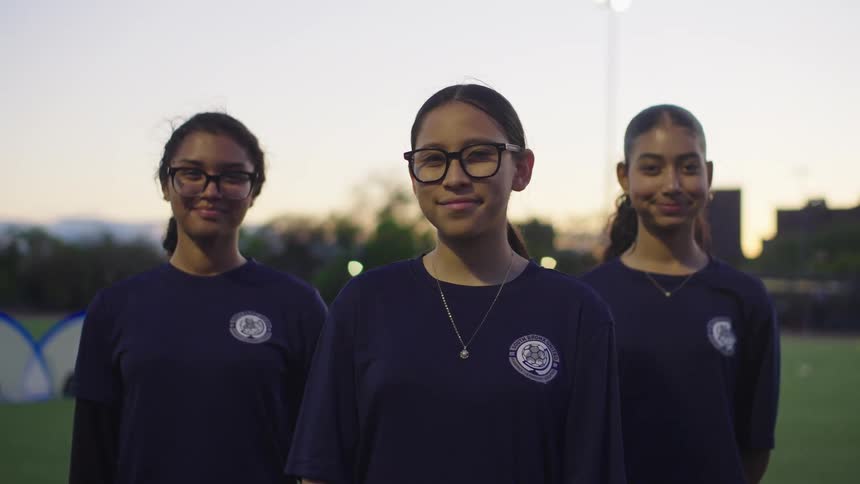 A slow-motion portrait of three young female soccer players smiling proudly on a city field at dusk
