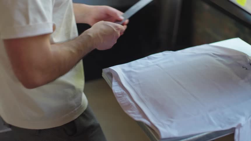 A man carefully prepares a white t-shirt for printing, using a ruler to ensure precise placement on the heat press machine in a workshop