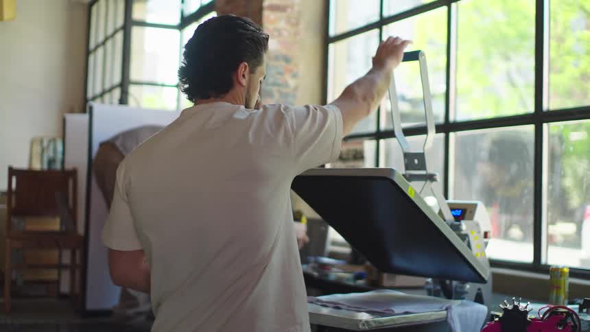 A skilled artisan operates a heat press machine in a sunlit creative workshop, applying a design to fabric