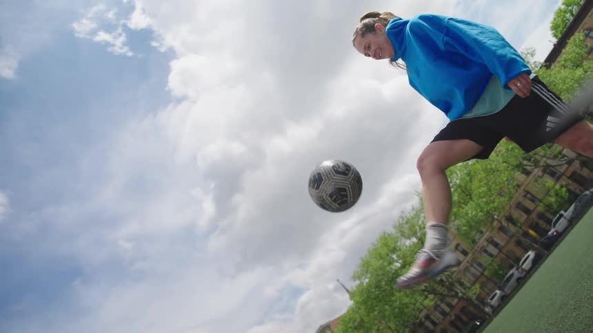 A low-angle, slow-motion shot captures a skilled female soccer player juggling a ball on an urban field under a partly cloudy sky