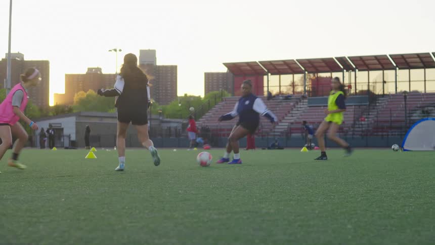 A women's soccer team practices running drills and playing on an urban field with Yankee Stadium in the background at sunset