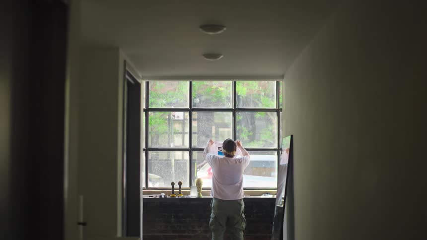 A creative man inspects a graphic design poster by holding it up to the natural light from a large window in his apartment hallway