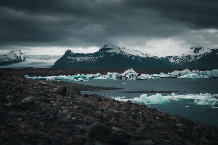 A dramatic and moody landscape of a glacial lagoon with floating icebergs and snow-covered mountains under a stormy sky