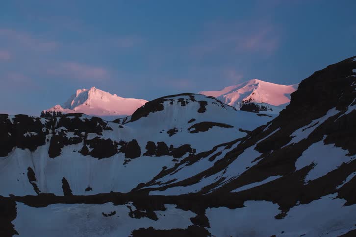 A stunning timelapse captures the pink alpenglow of sunset fading over a majestic, snowy mountain range as day turns to night