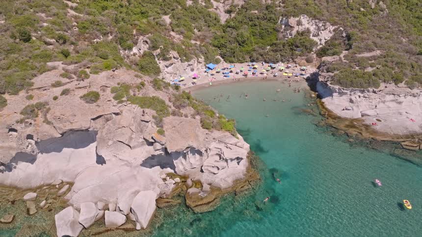 Stunning aerial shot pulling back to reveal a hidden beach cove where people enjoy the clear turquoise water on a sunny day