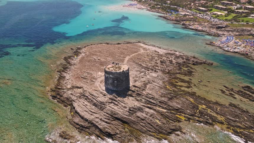 Stunning aerial footage rises over an ancient coastal tower on a rocky island, revealing a spectacular turquoise bay and bustling summer beach in Sardinia, Italy
