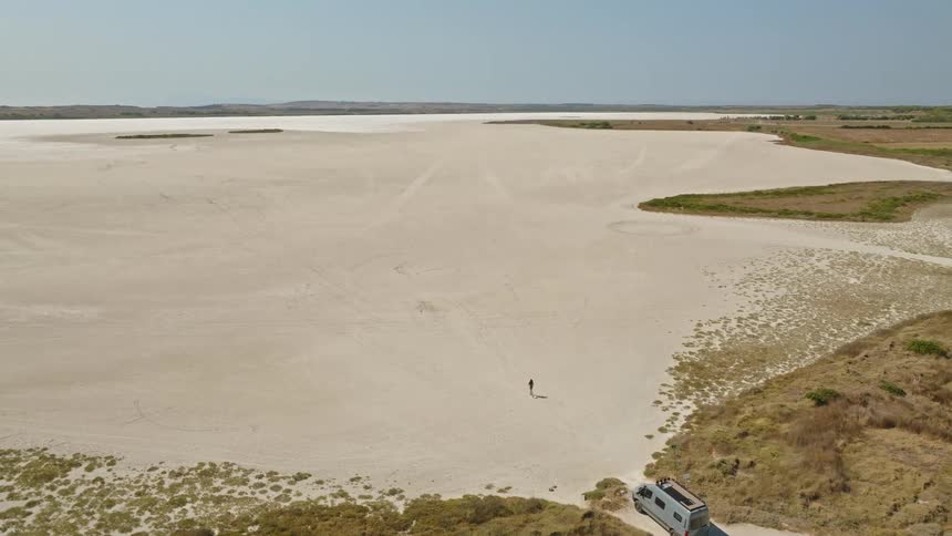 A lone person walks from their camper van into a vast, dry salt flat on a sunny day
