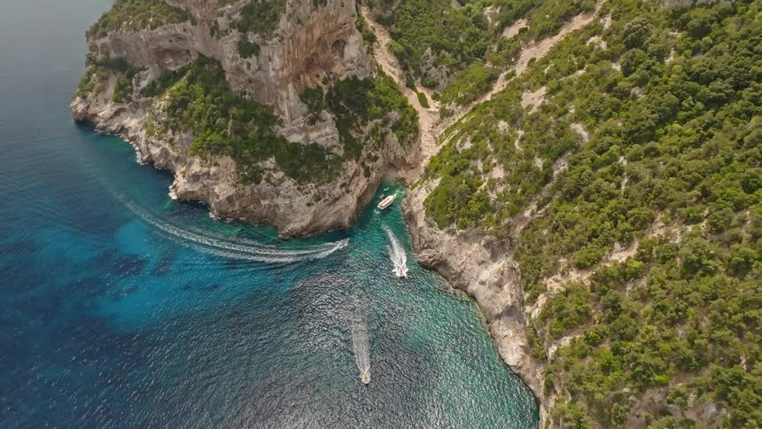 An epic aerial view of a dramatic, verdant coastline where boats explore a hidden turquoise cove nestled between massive cliffs