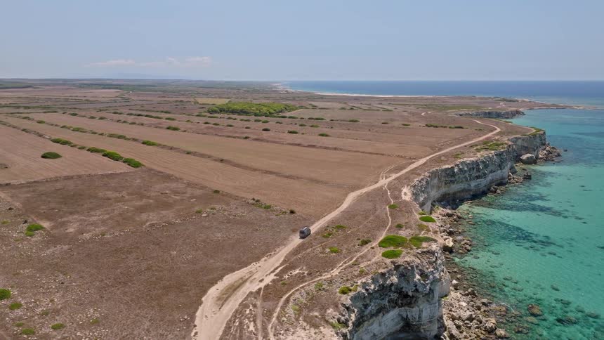 Aerial footage follows a lone vehicle on a dirt track along a dramatic cliffside coastline with vibrant turquoise waters