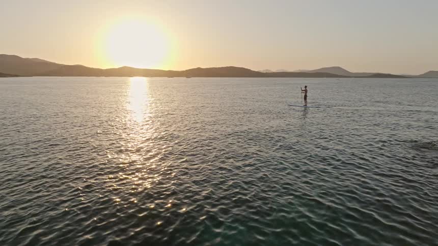 Aerial shot flies over a rocky coastline to reveal a man on a stand-up paddleboard enjoying the serene ocean at sunset