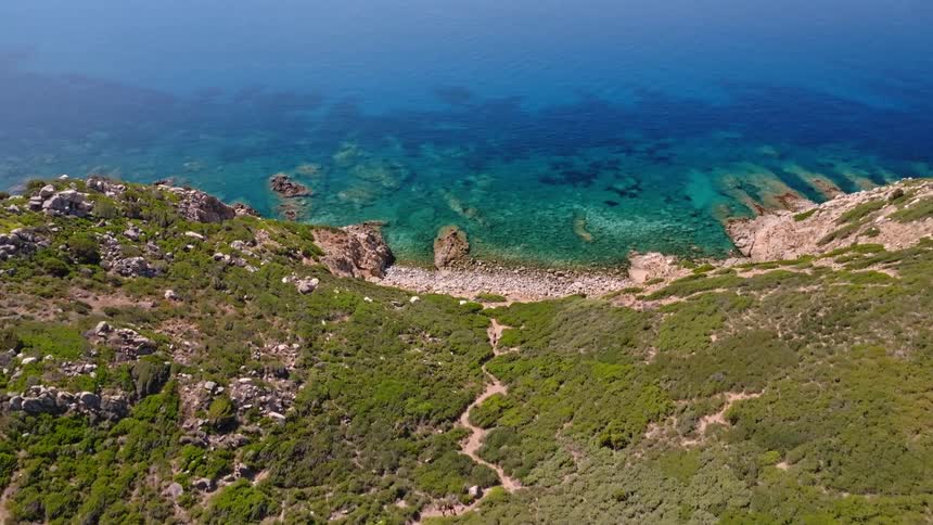 An epic aerial reveal of a winding coastal trail leading down a lush green hillside to a stunning secluded cove with crystal-clear turquoise water