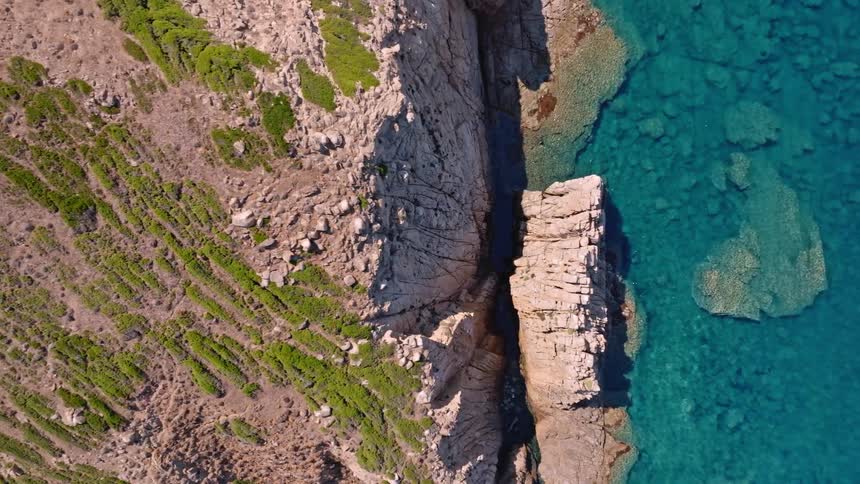 An aerial top-down view slowly pans over a dramatic rocky cliffside adjoining a calm, crystal-clear turquoise ocean