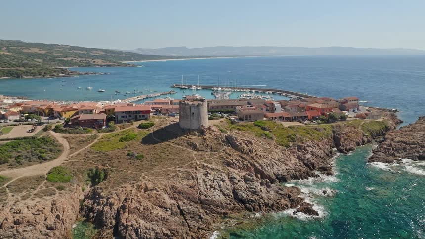 Ascending aerial shot reveals a historic stone tower on a rocky peninsula next to a picturesque coastal town and marina