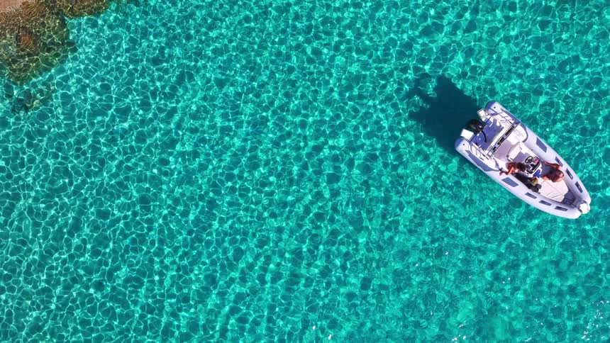 A peaceful aerial top-down cinemagraph captures a white boat anchored in the shimmering, transparent turquoise waters of a tranquil bay