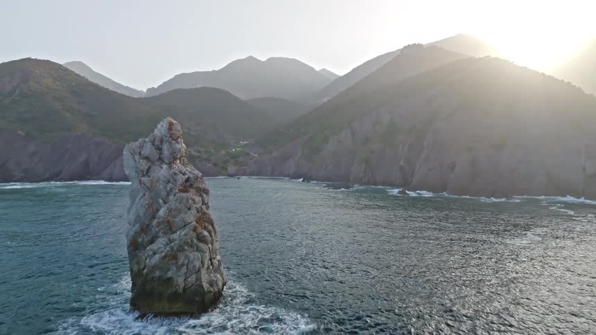 Aerial view pulls back from a rugged coastline to reveal a towering sea stack as the sun rises over the mountains