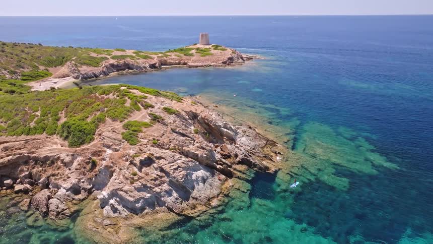 An aerial view shows a person on a paddleboard in crystal clear turquoise water next to a rocky coast with an ancient tower