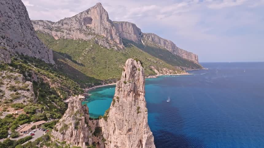 A breathtaking aerial shot flies over a majestic sea stack towards a hidden turquoise cove along a rugged, mountainous coastline