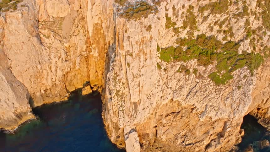 A stunning top-down aerial shot slowly pulls back to reveal a rugged, golden cliff dropping into the deep blue sea at sunset
