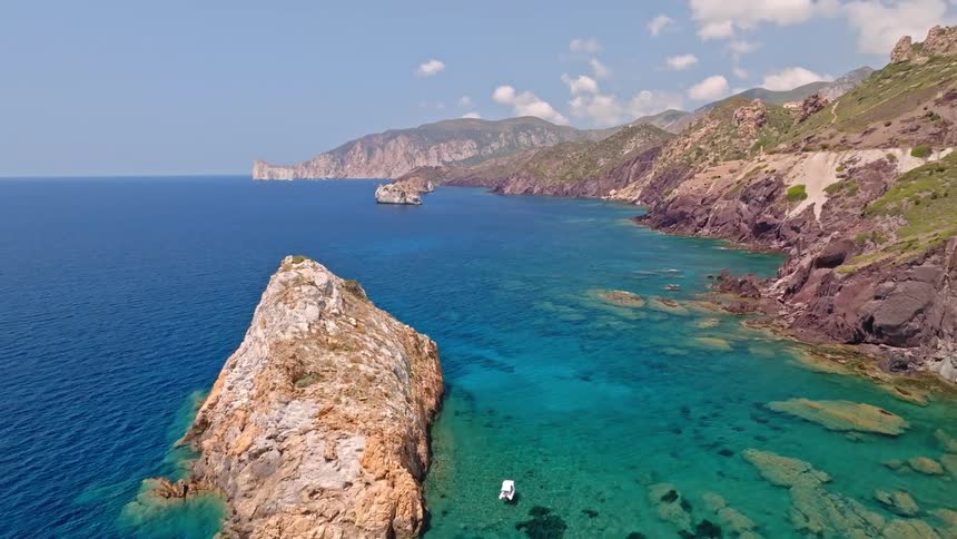An aerial drone flies backward revealing the stunningly clear turquoise and blue waters along a rugged and mountainous coastline on a sunny day