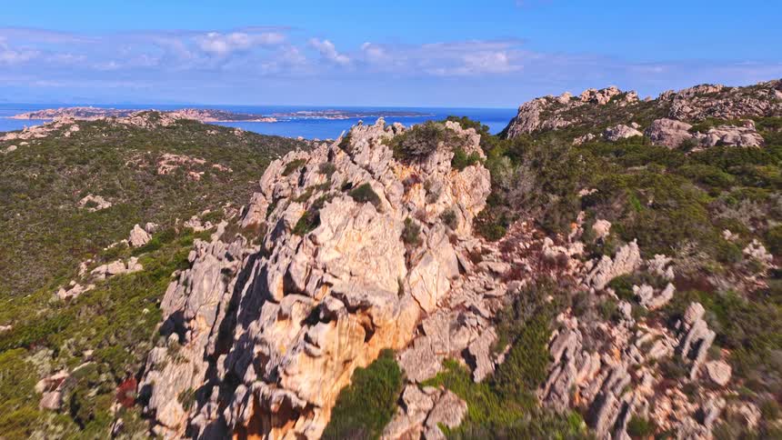 A scenic aerial shot flies over a rocky, scrub-covered mountain landscape towards the beautiful blue Mediterranean Sea